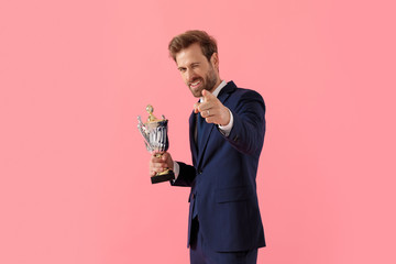 Charming businessman winking and pointing forward, smiling and holding trophy