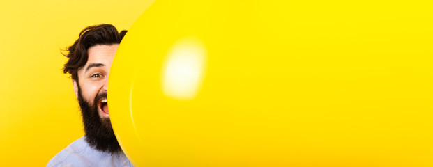 Handsome bearded man smiling and looking at camera while peeking out from above yellow balloon on yellow background, concept positive emotions, panoramic mock-up with space for text