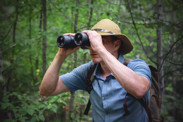 Tourist man is looking through binoculars in his hands on green forest background.