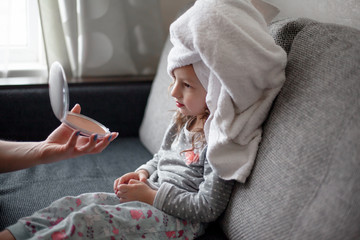 Beauty kid dressed pajamas and towel sit on the sof in the room. Close-up of woman hands holding the cosmetics and show to mirrow.