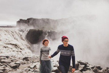 Obraz premium Young stylish couple in love walking together near famous Icelandic Dettifoss waterfall. Traditional wool sweaters, hat, red hair, gray skirt. Dramatic nordic landscape, cold weather in Iceland
