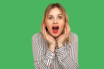 What a surprise! Excited blond girl in striped blouse standing with wide open mouth and raising hands in amazement, shocked by unbelievable success. indoor studio shot isolated on green background