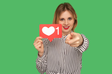 Hey you, put like to content! Pretty girl with red lips in glamour blouse holding social media heart icon and pointing to camera, recommending to follow blog. studio shot isolated on green background