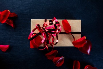 Valentine's Day, love. Red roses petals, heart and gift with a red ribbon on a black background
