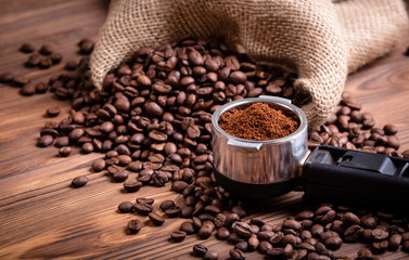 Roasted coffee beans waking up from a jute coffee bag. A porta filter with ground coffee on a brown vintage wooden background.