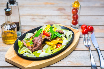 Fried pieces of meat with vegetables in a hot pan on a wooden table in a restaurant.