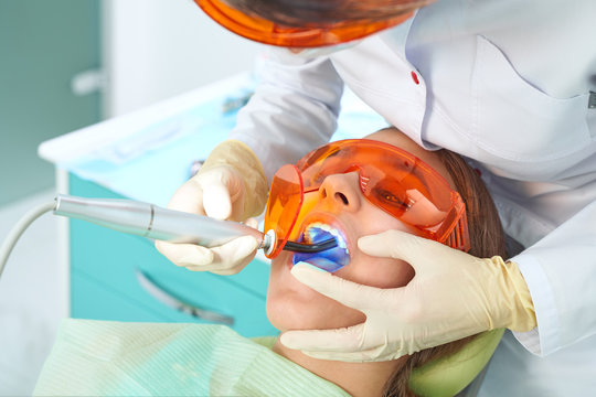 Girl Child At The Doctor. Dentist Places A Filling On A Tooth With Dental Polymerization Lamp In Oral Cavity. Over Clinic Background