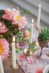 A bouquet of blooming peony, roses and other flowers in a vase on a rustic table surrounded by candles with vintage candlesticks. Boho chic for the holiday. Scenery decoration for dinner.