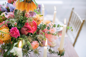A bouquet of blooming peony, roses and other flowers in a vase on a rustic table surrounded by candles with vintage candlesticks. Boho chic for the holiday. Scenery decoration for dinner.