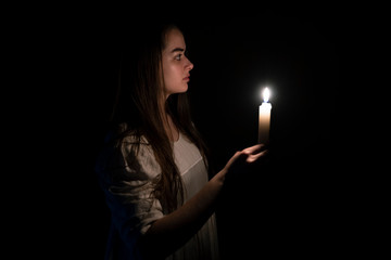 A young girl in an old white dress holding a candle in her hand. Side view. Dark background. Scary horror concept. 