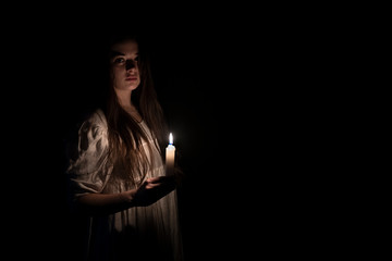 A young girl in an old white dress holding a candle in her hand and staring at the camera. Copy space on the right side. Dark background. Scary horror concept. 