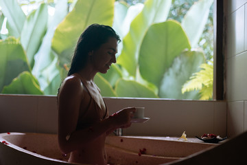 Woman sitting in stone bath tube with tropical flowers and drink herbal tea with jungle view, organic skin care, luxury spa hotel, Bali Indonesia