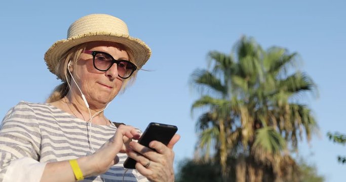 Blonde elderly woman in sunglasses and straw hat choosing a song on mobile phone.