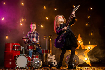 Kids pretending to be in a rock band and play and sing at studio or stage. Girl play on guitar and boy on drums.