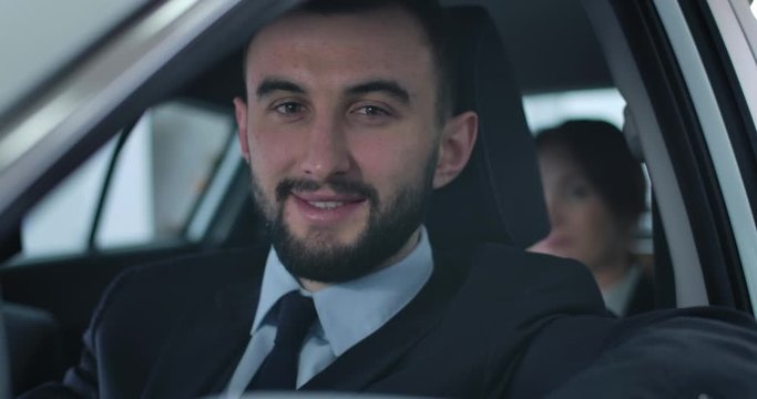 Close-up face of young handsome Caucasian man looking at camera and smiling. Professional chauffeur sitting on driver's seat in car with passenger at the background. Cinema 4k ProRes HQ.