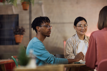 Multi-ethnic group of three young people working on project together while sitting at table in cafe, copy space