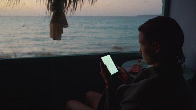 girl reading ebook reader inside a hut in front of a caribbean beach during sunset