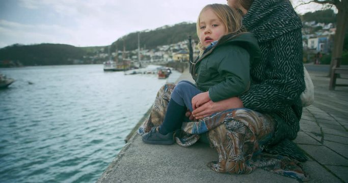 Preschooler sitting by river in seaside town with his mother