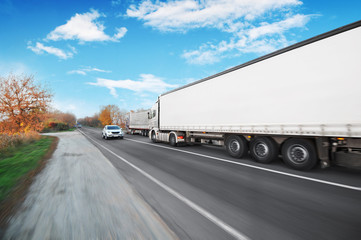 A big truck and trailer with other vehicles driving fast on the countryside road against a blue sky with clouds