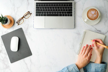 Flat lay workspace. Woman hand with coffee cup, smartphone, computer, notebook, planner and stationary with copy space on marble table background. Top view.