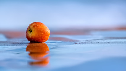 Red apple lying on a sandy beach in shallow surf with reflections in the blue water and with the ocean as a background