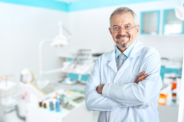 Naklejka na ściany i meble Portrait of glad smiling doctor in white uniform standing with crossed hands on blue dental clinic background