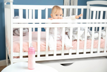 Cute baby sitting in crib near bottle of milk on table