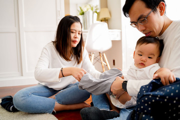 Young Asian father sitting on floor and holding infant boy while mother holding baby clothing and together taking care of son and changing diapers