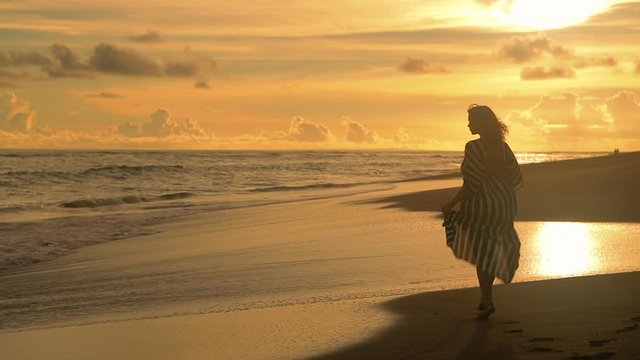 Young sexy woman in black dress walking and looking at the sunset at the sand beach. Pretty girl walking towards ocean. Femininity concept. City people at vacation. Business woman at summer vacation.