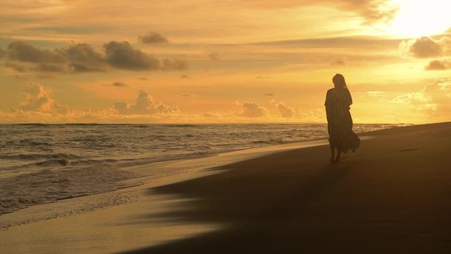 Young sexy woman in black dress walking and looking at the sunset at the sand beach. Pretty girl walking towards ocean. Femininity concept. City people at vacation. Business woman at summer vacation.