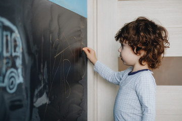 Fototapeta na wymiar Curly little boy draws chalk on a board in his room