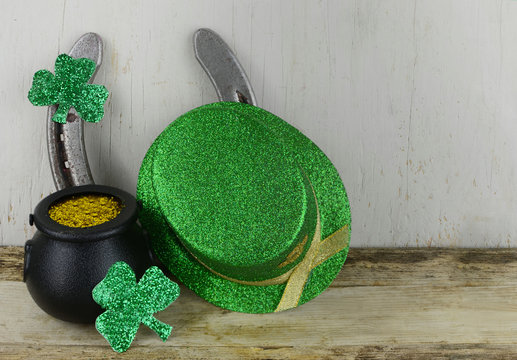 Saint Patrick's Day image of glittered green sharmocks, a horseshoe, a pot of gold dust and a leprechaun's hat on a rustic wooden background.