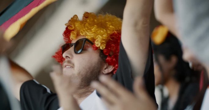German soccer supporters cheering their national team from stadium. Man wearing a colorful wig and sunglasses having fun while watching a game from fan zone.