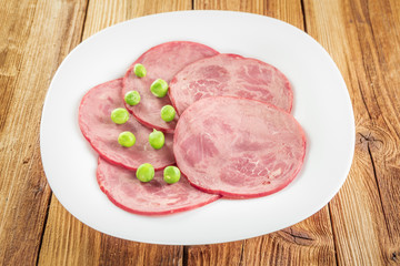Slices of sliced ham and green peas on a white plate on a wooden table.