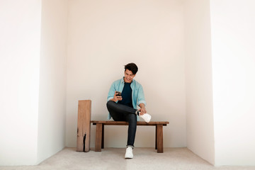 Happy Young Man in Casual wear Using Mobile Phone while Sitting on Bench by the Wall. Lifestyle of Modern People. Wide Shot, Full Length