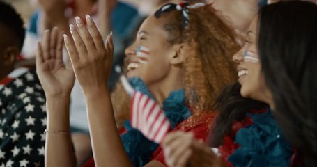 Women with USA flag sitting in stadium stands clapping and cheering for their team. Happy USA soccer fans enjoying watching a match in stadium. - Powered by Adobe