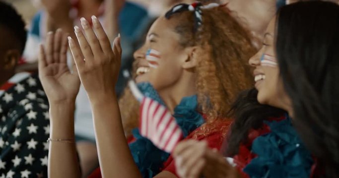 Women with USA flag sitting in stadium stands clapping and cheering for their team. Happy USA soccer fans enjoying watching a match in stadium.