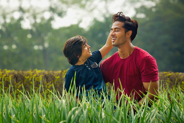 Smiling young boy sitting with his father in garden and touching his hair with grass in the foreground.