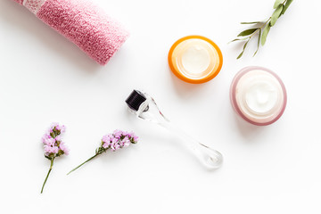 Cosmetologist tools. Dermaroller, creams near leaves and flowers on white background top-down flat...