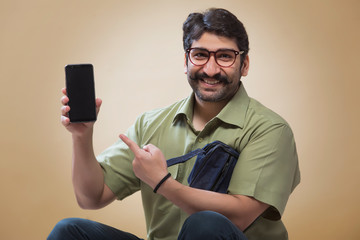 Smiling man wearing eyeglasses pointing towards a mobile phone which is held in other hand.