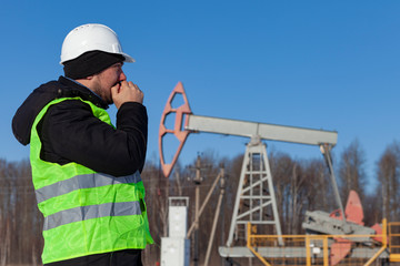Oil worker in orange uniform and helmet warms hands from the cold on of background the pump sucks and blue sky