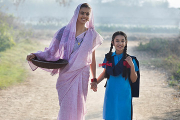 Female farmer or rural woman walking on village street along with her school going daughter carrying an iron gold pan.