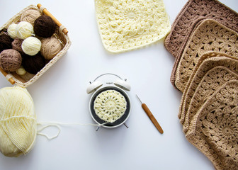 Crochet on a white background. On a white table are balls of yarn, square motifs, an alarm clock, a hook. Knitting / hobby time concept.