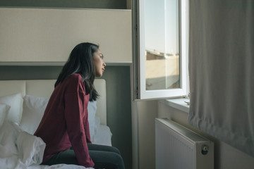 Asian woman is sitting on the bed and looking out to the window.