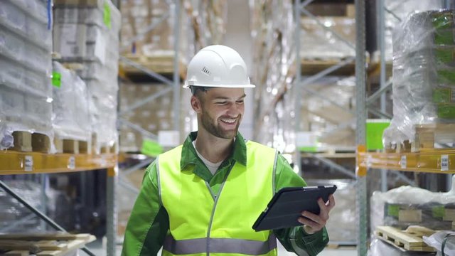 man warehouse worker walks through rows of storage racks with merchandise and looks at digital tablet. male smiling. architect walking through logistics shelves of factory. male in storage with goods