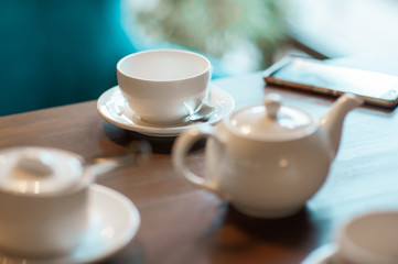 White cups and a white teapot on a table in a cafe. A mobile phone lies next to the cup.