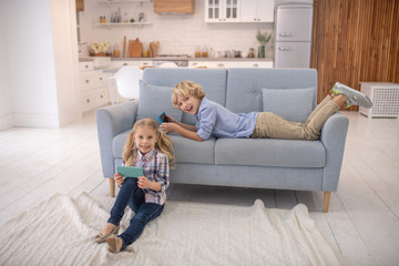 Boy lying on sofa, girl sitting on the floor with gadgets