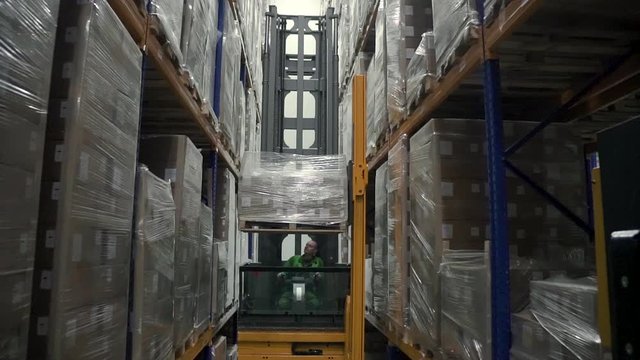 Young man is lifting cargo using forklift in industrial warehouse during working day. Caucasian male worker works while sitting in loading machine, puts goods on high shelves of storehouse in factory
