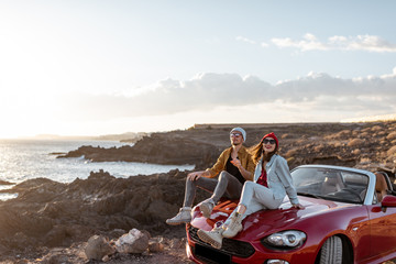 Young lovely couple enjoying landscapes, sitting together on a car hood, traveling by car on the rocky ocean coast. Carefree lifestyle, love and travel concept