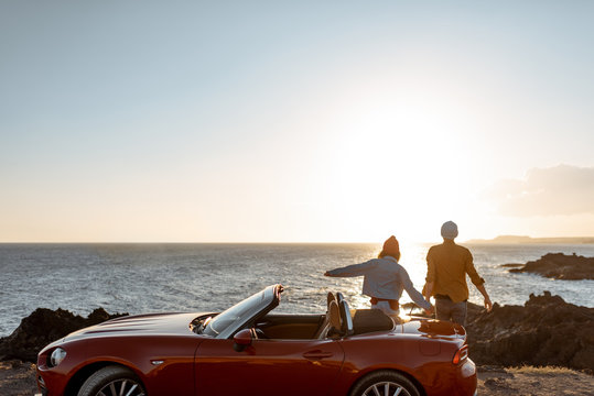 Couple enjoying beautiful views on the ocean, standing together near the car on the rocky coast, wide view from the side with copy space